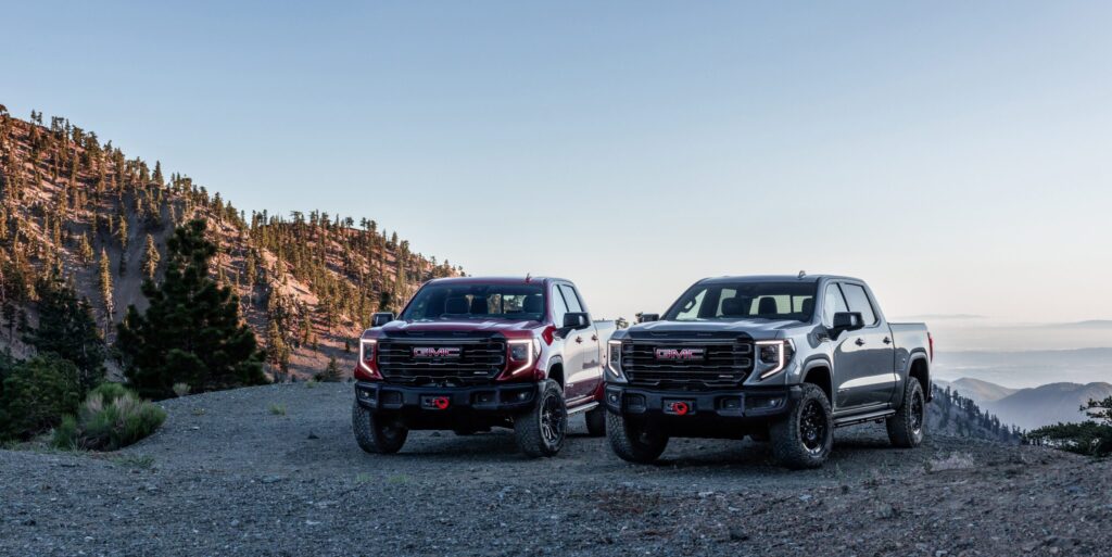 Papel de parede de duas picapes GMC Sierra AT4X, uma vermelha e uma cinza-chumbo, estacionadas em uma trilha de montanha rochosa.