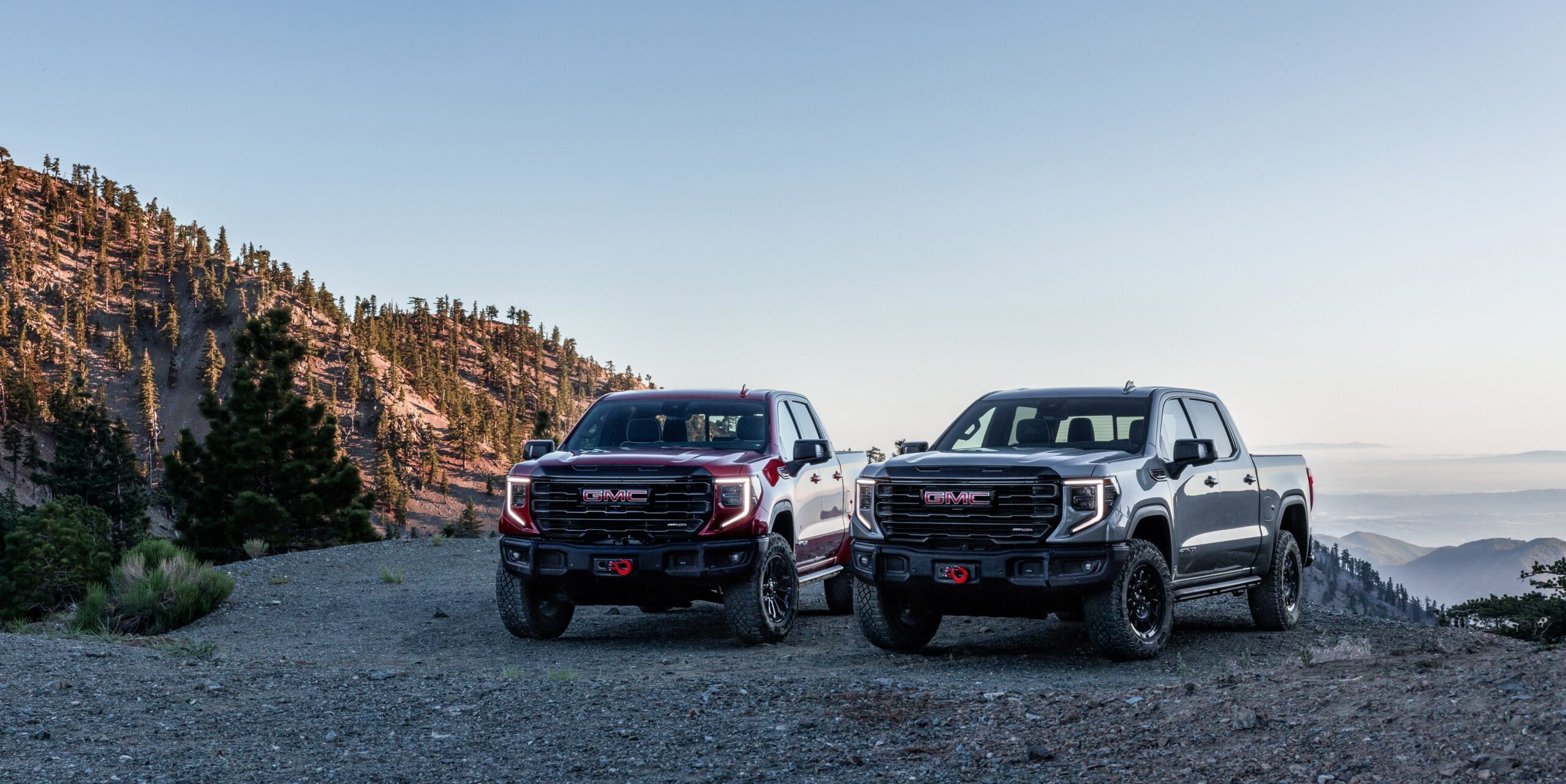 GMC Sierra AT4X Papel de parede de duas picapes GMC Sierra AT4X, uma vermelha e uma cinza-chumbo, estacionadas em uma trilha de montanha rochosa.