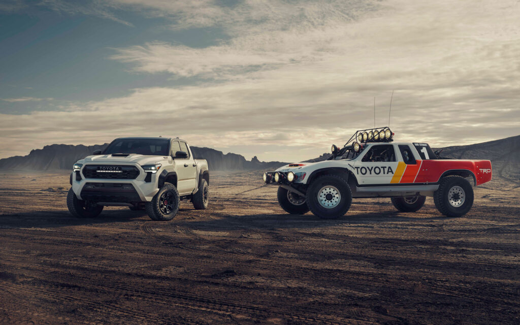 Fundo de tela com a nova picape Toyota Tacoma 2024 branca ao lado de uma picape de corrida clássica da Toyota TRD (Baja truck) no deserto.