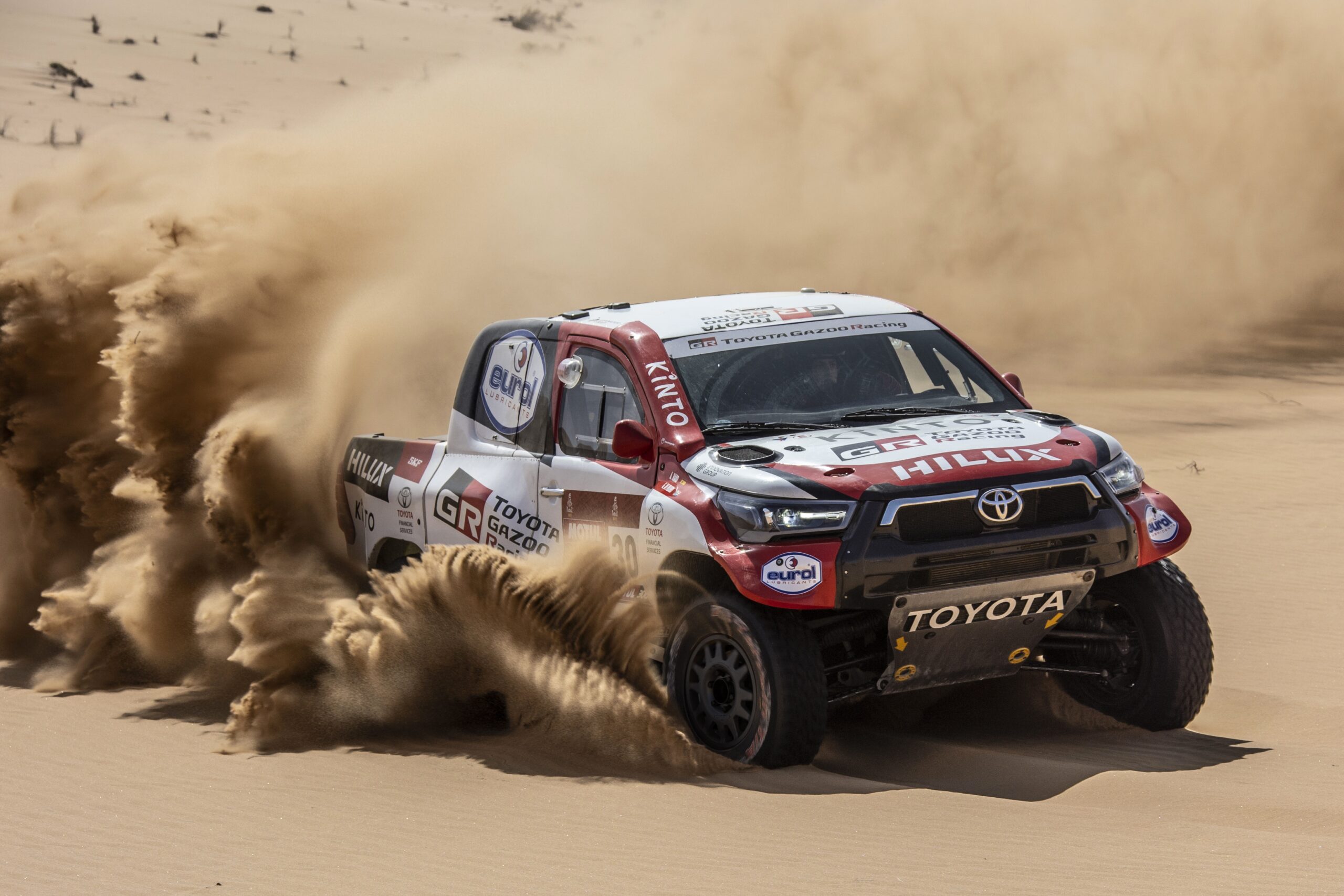 Toyota Hilux GR DKR (Rally Dakar) Fundo de tela do Toyota Hilux GR DKR (Rally Dakar) com a pintura da Gazoo Racing, correndo em alta velocidade e levantando uma nuvem de areia no deserto.