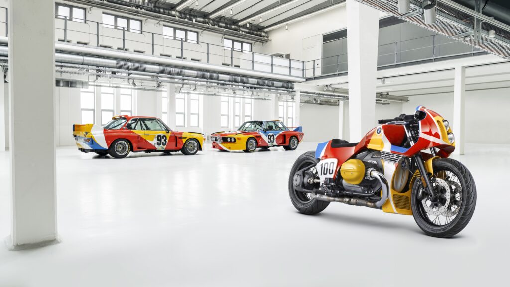 Fundo de tela com dois carros de corrida clássicos BMW 3.0 CSL "Art Car" (Calder e Stella) e uma moto BMW R nineT customizada com a mesma pintura, em um estúdio branco.