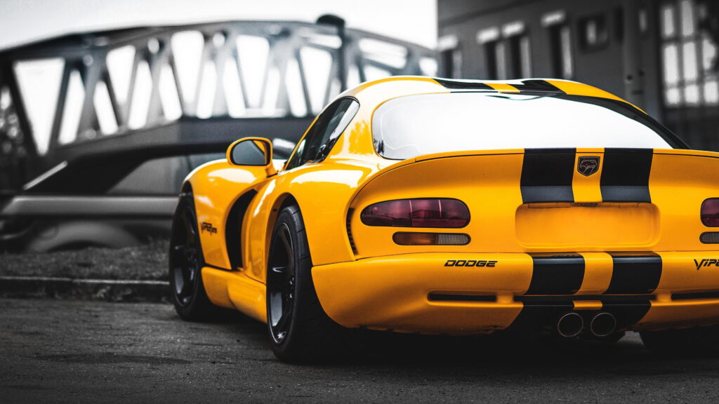 Papel de parede de um Dodge Viper GTS amarelo com listras pretas, visto de trás, com um fundo industrial/ponte desfocado.