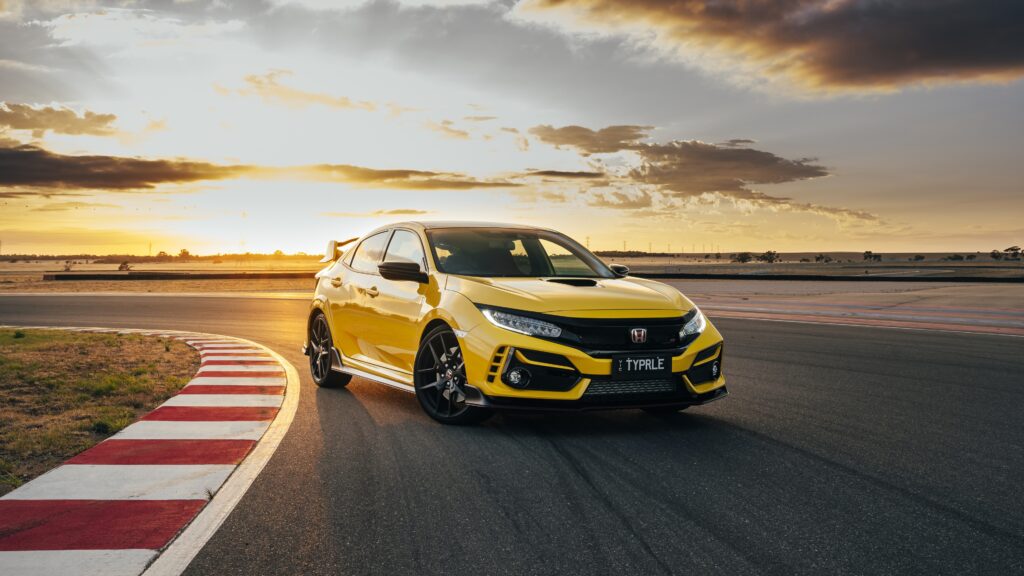 Papel de parede de um Honda Civic Type R (FK8) amarelo (Sunlight Yellow) em uma pista de corrida, fazendo uma curva sobre a zebra durante o pôr do sol.