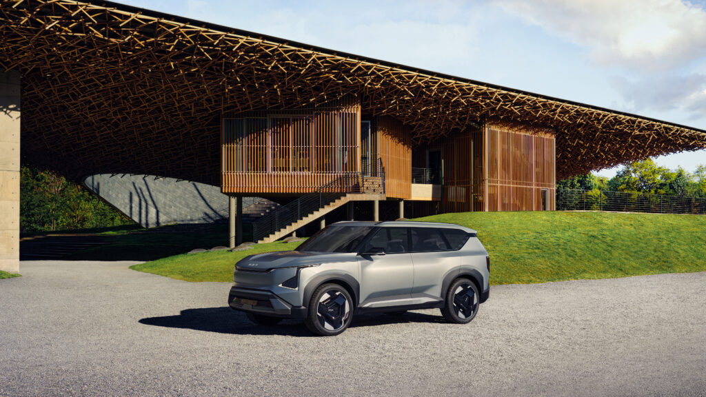 Fundo de tela do SUV elétrico Kia EV9 na cor prata, estacionado em frente a um prédio de arquitetura moderna feita de bambu.