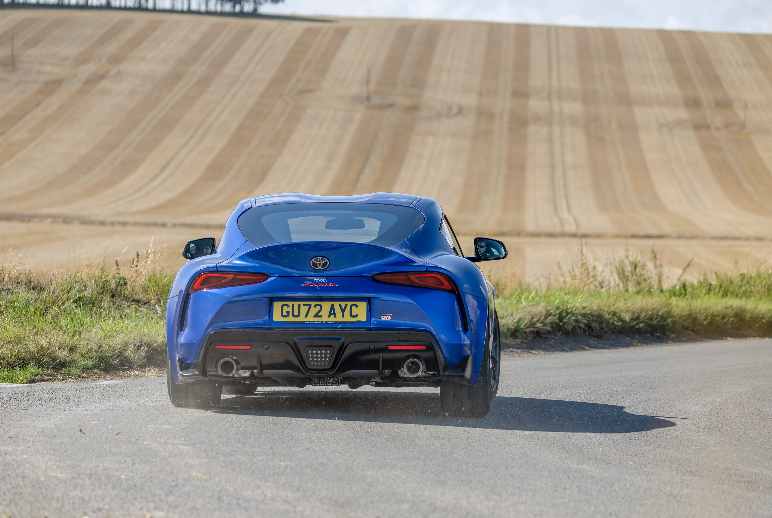 Toyota GR Supra Papel de parede da traseira do Toyota GR Supra azul (A90) em movimento em uma estrada de campo, com um campo de trigo ao fundo.