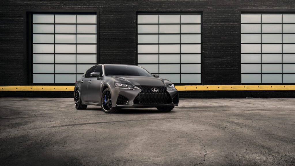 Papel de parede de um sedan esportivo Lexus GS F na cor cinza fosco (Nebula Gray), estacionado em um pátio industrial com janelas ao fundo.