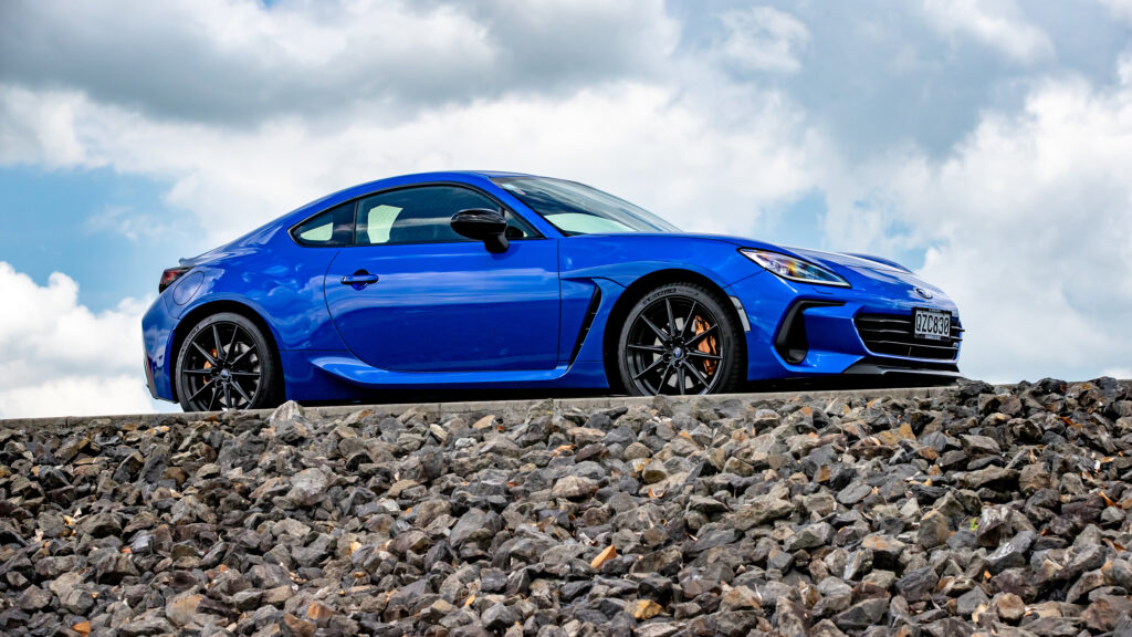 Fundo de tela do novo Subaru BRZ azul (World Rally Blue), visto de perfil estacionado sobre uma mureta de pedras, com céu azul e nuvens ao fundo.